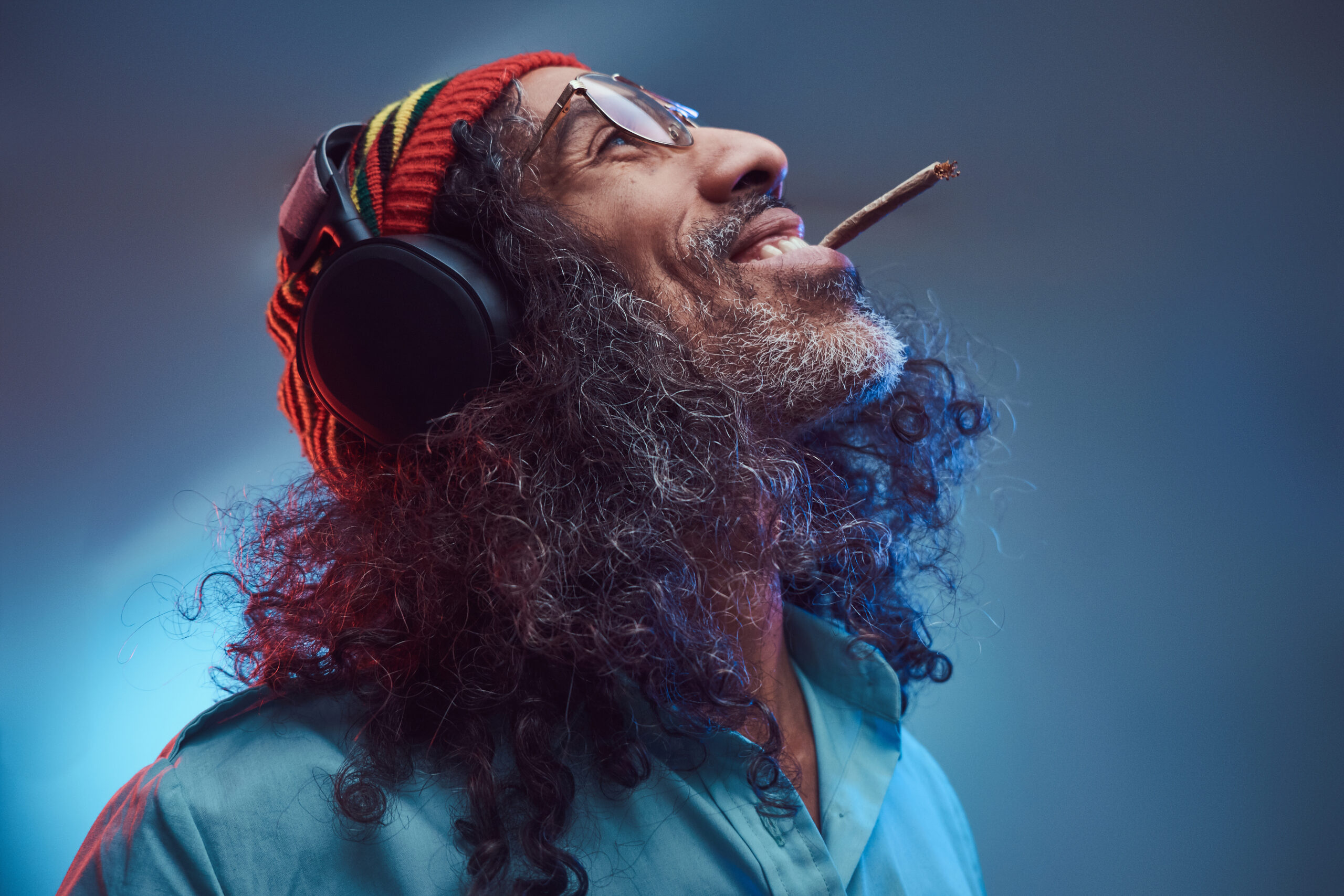 Studio portrait of African Rastafarian male enjoys music in headphones and smoking weed. Isolated on a blue background.